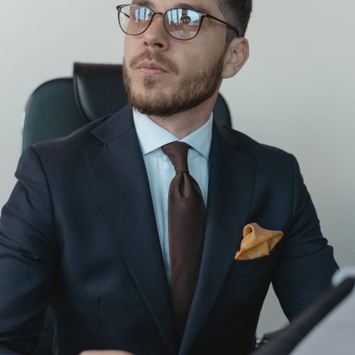 Young professional businessman wearing a suit and glasses, sitting at a desk with documents.